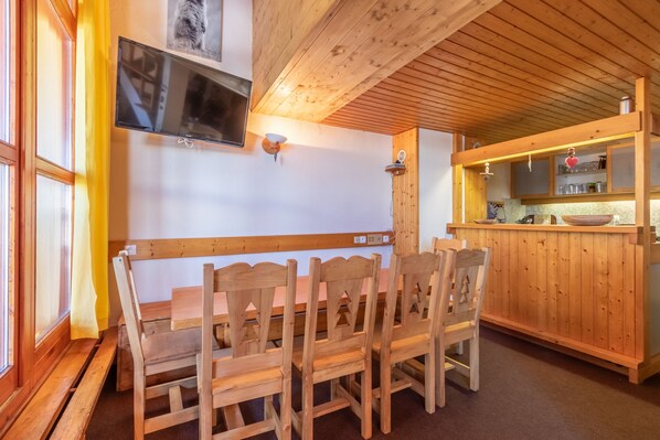 Ein Essbereich mit Holztisch und -stühlen, einem an der Wand befestigten Fernseher und einer Küchenzeile mit Holzausstattung und Regalen.