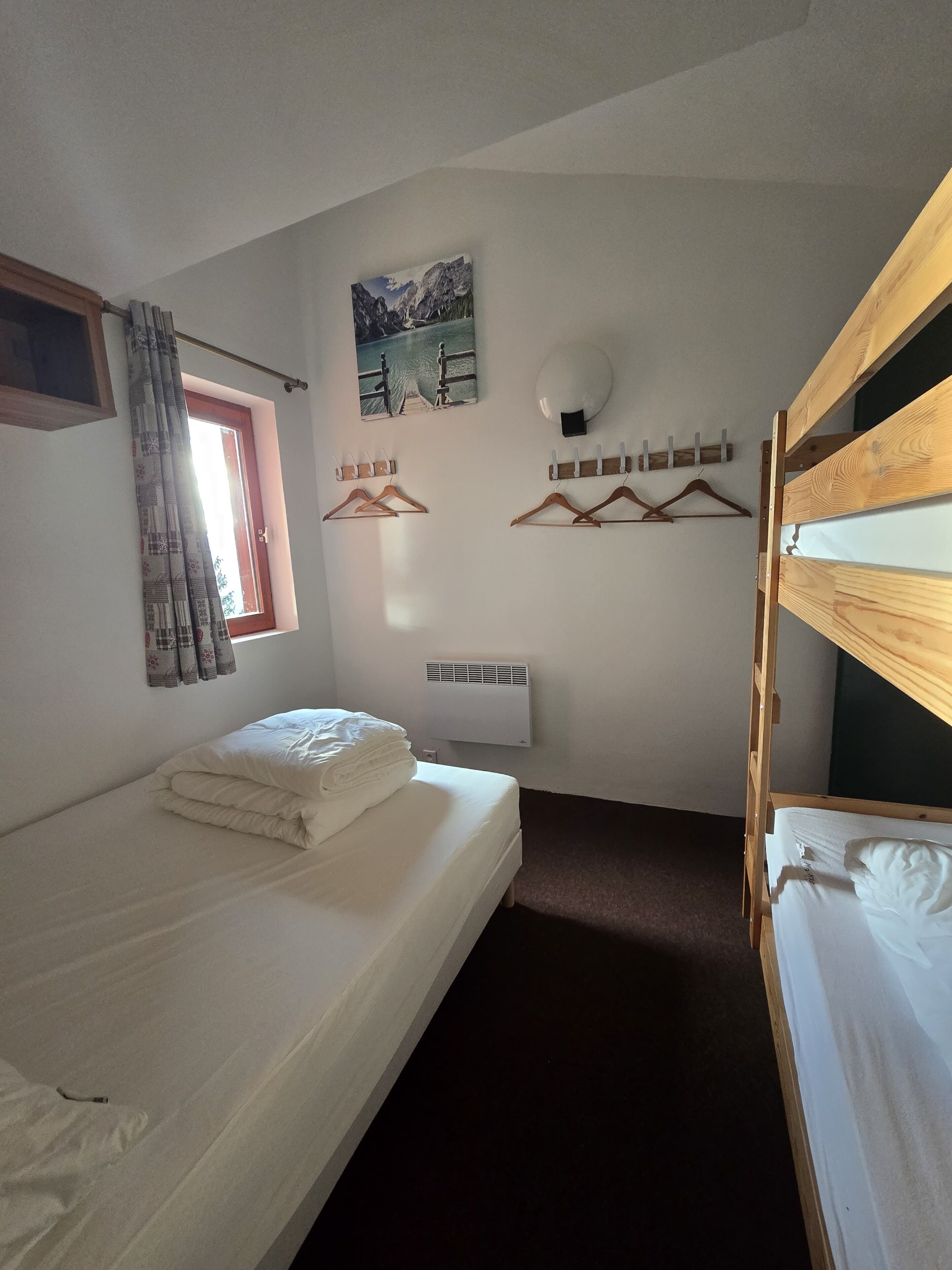 A room with a bunk bed, a single bed, a window with curtains, a wall-mounted heater, and a painting.