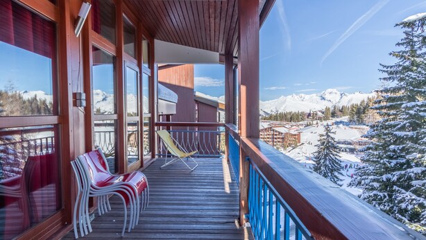 Une terrasse en bois avec des chaises, une vue sur des montagnes enneigées et une piste de ski en arrière-plan.