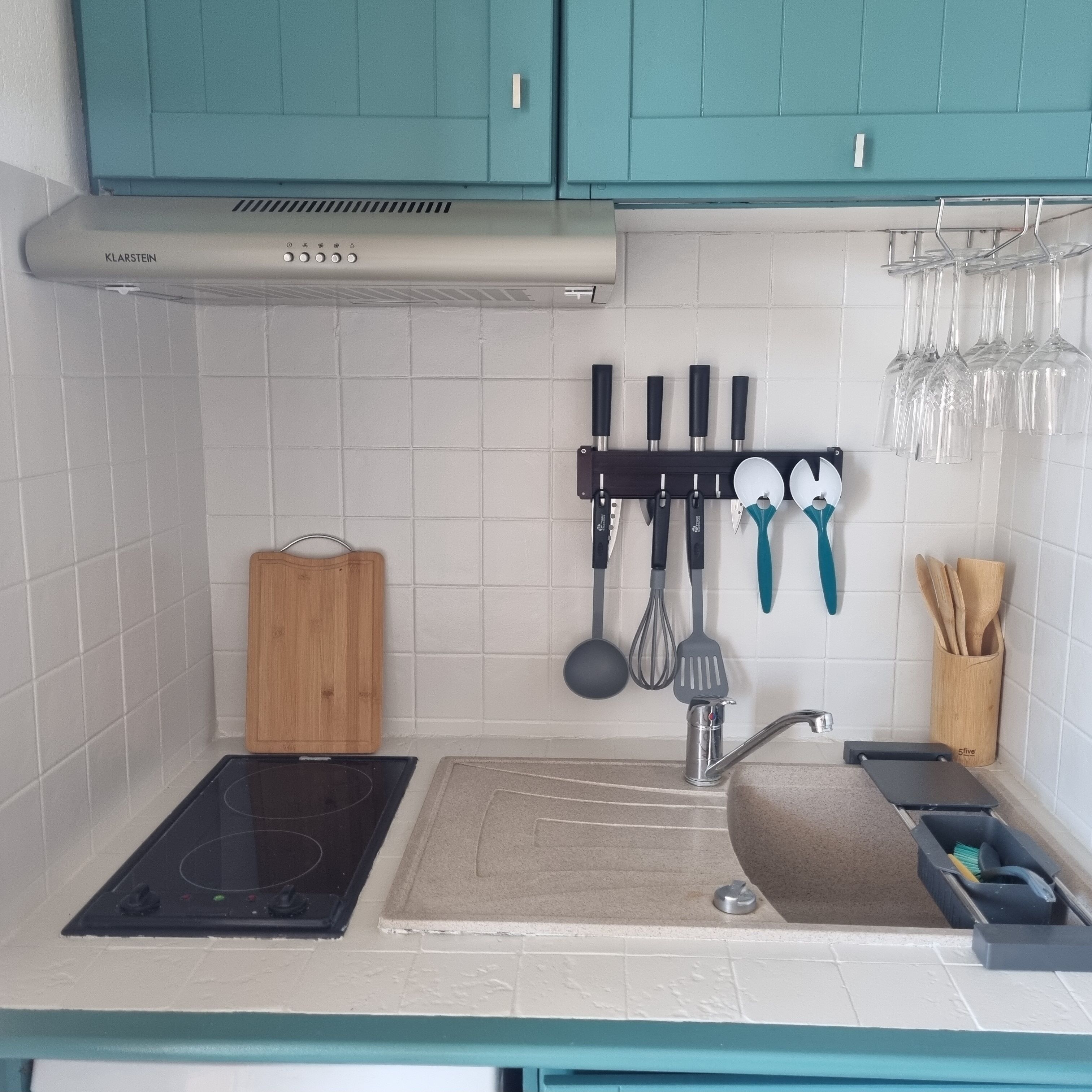 A kitchen with teal cabinets, a gas stove, a sink with a cutting board, and utensils hanging above the sink.