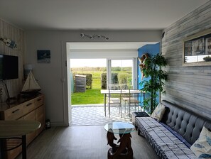 Ein modernes Wohnzimmer mit einer Couch, einem Glastisch und einem Fernsehgerät auf einer Holzkommode. Durch eine Glastür ist ein Garten und ein kleines Gartenhaus zu sehen.