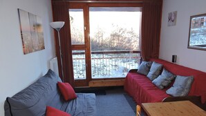 A cozy room with a grey sofa, a red sofa, a wooden table, and a large window with a view of a snowy landscape. - Studio Arc 2000, 4/5 persons, residence Bel Aval, Charlotte Perriand style. (Bourg-Saint-Maurice)