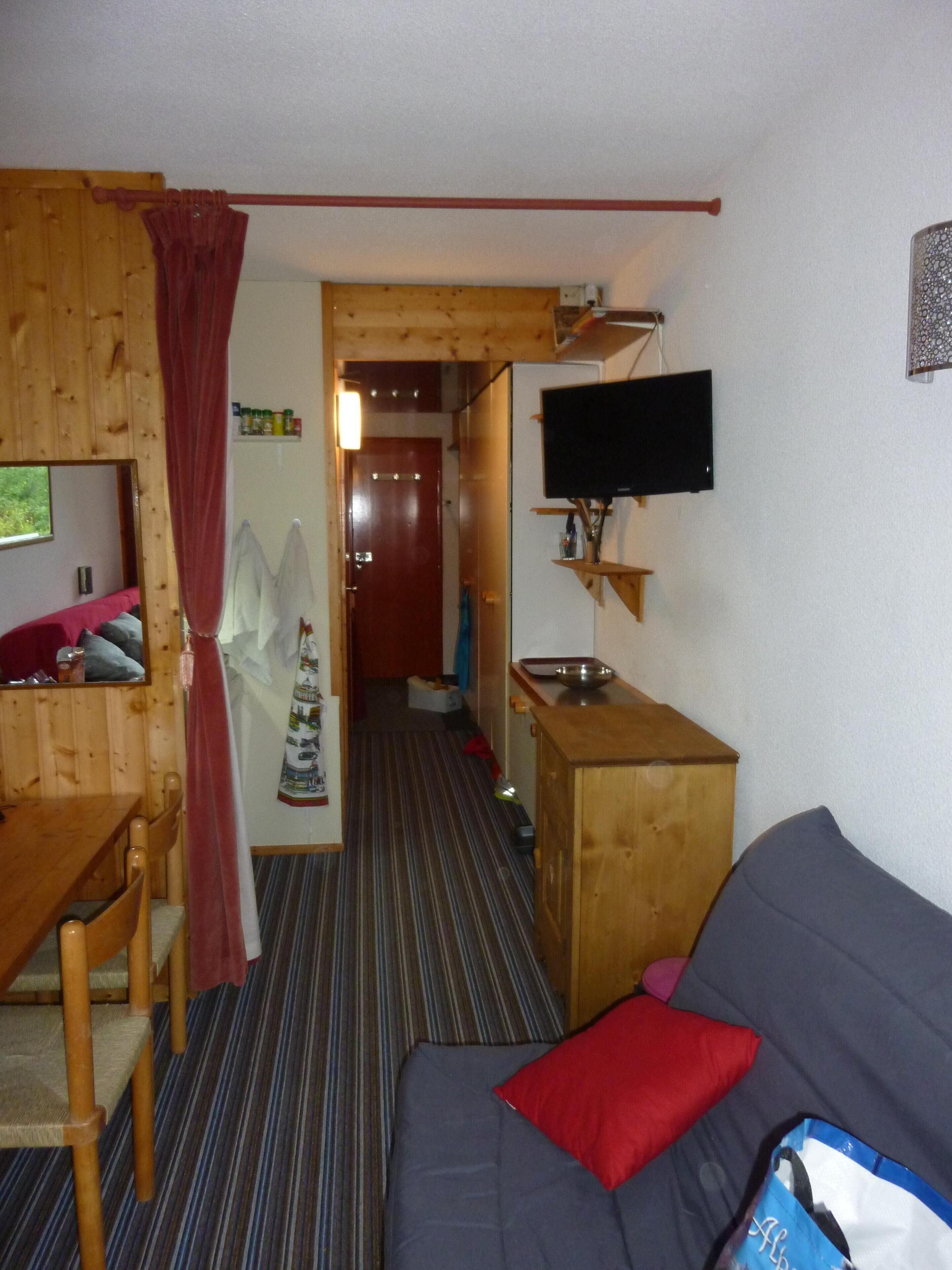 A room with a wooden dining table, a sofa, a television mounted on the wall, and a door leading to another room.