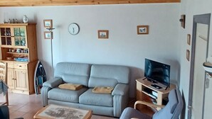 Un soggiorno con soffitto in legno, un divano grigio, un televisore su un mobile in legno e un tavolino con una cornice.