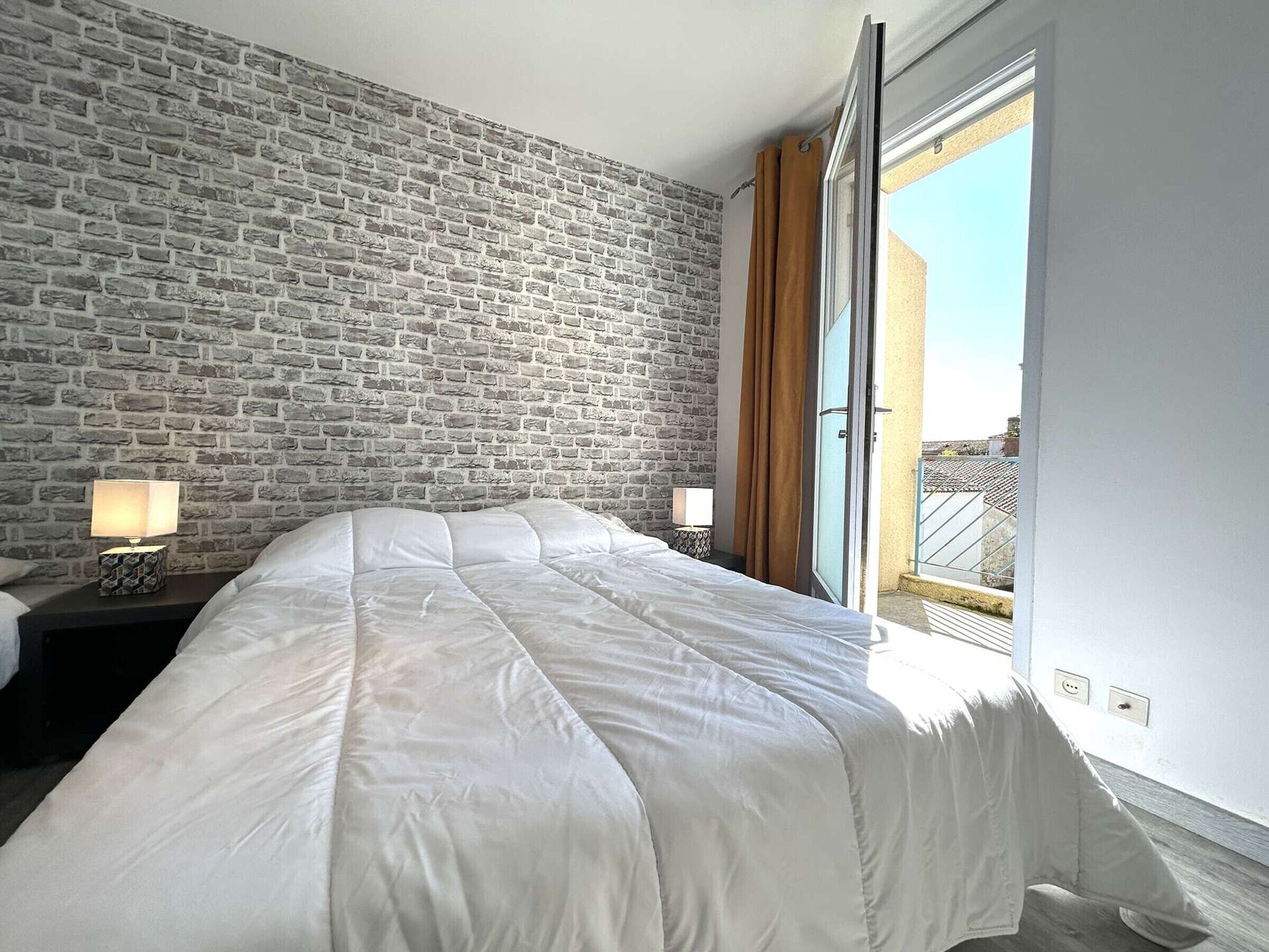 A bedroom with a brick wall, a bed with white bedding, two bedside tables with lamps, and a view of a balcony.