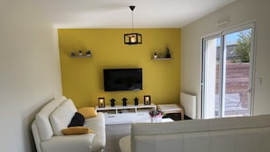 A living room with a white sofa, a white armchair, a TV mounted on the wall, and a small table with decorative items. - Pretty house with enclosed garden close to amenities in ERDEVEN. (Erdeven)