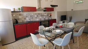 A compact kitchen with red cabinets, a modern stove, and a dining area set for four. - OMAHA BEACH COTTAGE BETWEEN EARTH AND SEA (Formigny)
