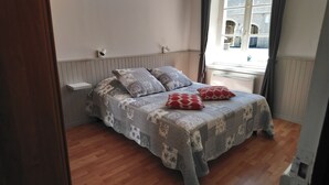 A bed with patterned bedding and two red pillows, a white shelf, and a window with a view of buildings. - OMAHA BEACH COTTAGE BETWEEN EARTH AND SEA (Formigny)
