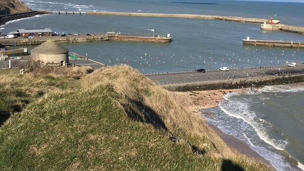 A coastal view with a grassy hill, a beach, and a pier extending into the sea. - OMAHA BEACH COTTAGE BETWEEN EARTH AND SEA (Formigny)