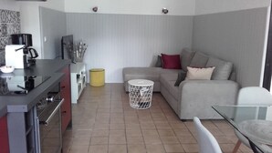 A compact living space with a kitchenette, a sofa, a small round table, and a trash bin. - OMAHA BEACH COTTAGE BETWEEN EARTH AND SEA (Formigny)