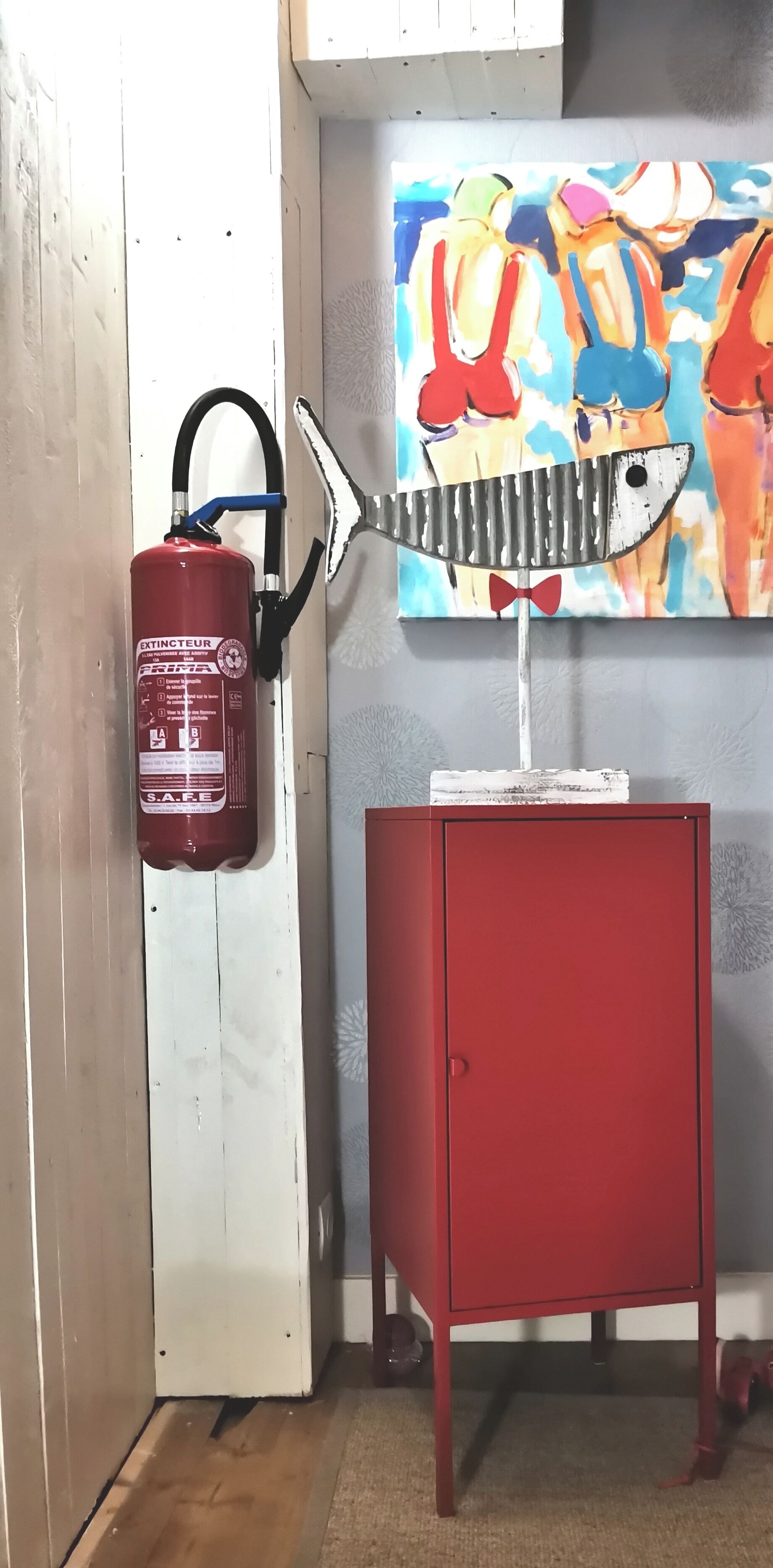 A red fire extinguisher mounted on a white door, a red cabinet, and a colorful abstract painting on the wall.