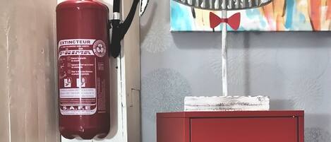 A red fire extinguisher mounted on a white door, a red cabinet, and a colorful abstract painting on the wall.
