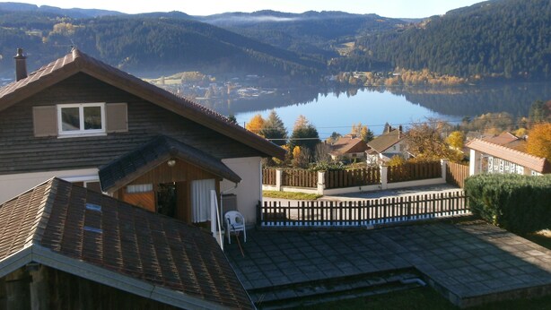 A house with a sloped roof, a wooden deck, and a view of a lake and mountains. - Cottage with magnificent view of the Lake in Gérardmer, 8 people (Gérardmer)