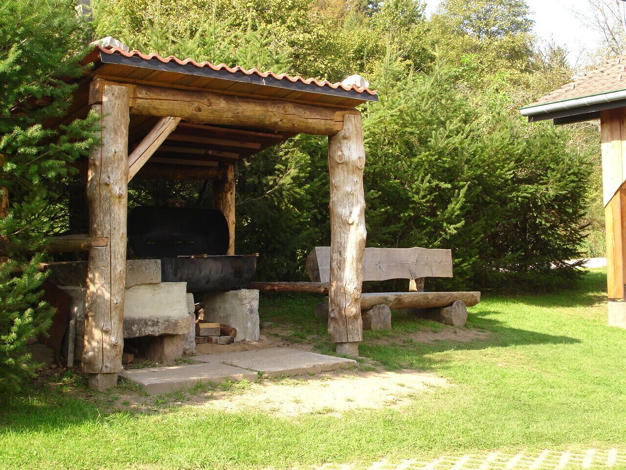 Eine rustikale Holzkonstruktion mit Ziegeldach, möglicherweise ein Pavillon oder Schutzhaus, umgeben von Grünpflanzen und einer Pflasterfläche.