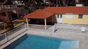 Uma piscina retangular com cerca branca, um espaço de estar coberto e um edifício com telhado de telhas vermelhas ao fundo.
