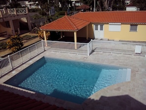 Une piscine rectangulaire entourée d’une clôture blanche, un patio couvert et, au fond, un bâtiment avec un toit recouvert de tuiles rouges.