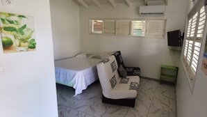 Quarto pequeno e bem iluminado com cama, sofá, televisão e janela com persianas.