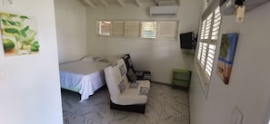 Une petite chambre bien éclairée, équipée d’un lit, d’un canapé, d’une télévision et d’une fenêtre avec des volets.