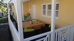 Un espace extérieur abrité, aménagé pour prendre les repas, avec une table pour deux personnes, une chaise rembourrée de couleur verte et une fenêtre avec des volets.