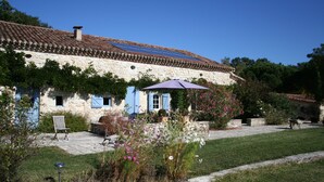 Un bâtiment en pierre avec un toit en tuiles, un jardin fleuri et une chaise à l’extérieur.
