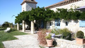 Un bâtiment en pierre doté d’une tour, d’un porche couvert et d’un jardin agrémenté de plantes en pot.
