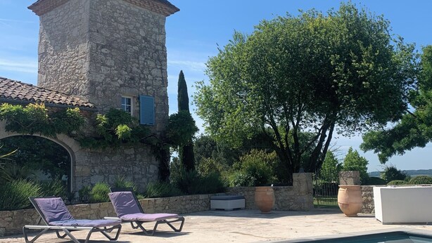Une tour en pierre surmontée d’un toit conique, une piscine avec des chaises longues et un grand arbre dans un cadre extérieur ensoleillé.