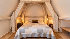 A bedroom with a sloped ceiling, wooden beams, a quilted bedspread, two bedside lamps, and a bench.