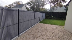 Una tanca de metall gris amb barres verticals, un camí de grava i un pràt verd.