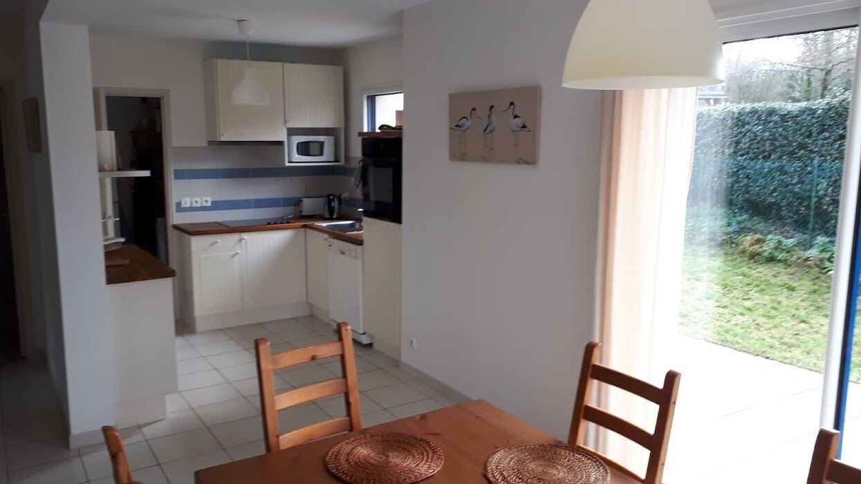 Une cuisine compacte avec des meubles blancs, un four à micro-ondes et un évier. On y trouve une table à manger avec des chaises, et une fenêtre donne sur un jardin.