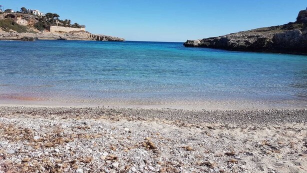Ein Strand mit klarem, blauem Wasser, felsigem Gelände und einem Gebäude links.