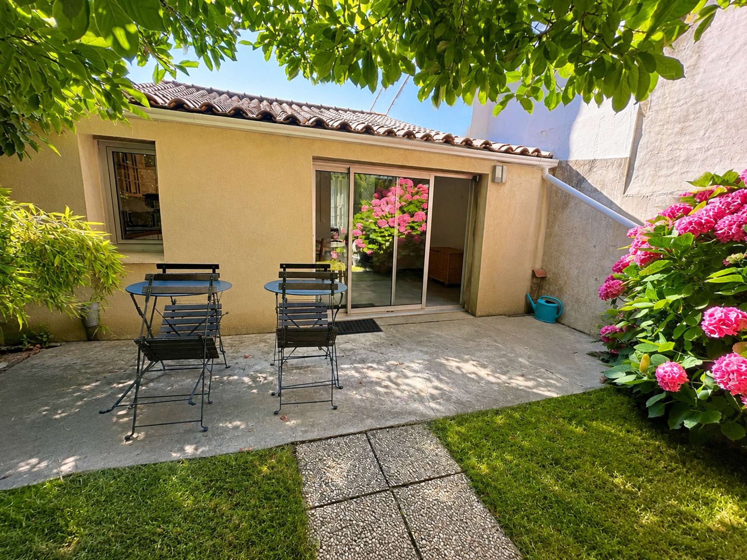 Une terrasse avec deux tables et des chaises, un petit jardin avec de la pelouse et des fleurs, et un bâtiment doté d’un toit en tuiles et d’une porte-fenêtre coulissante.