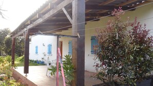 A covered patio area with a wooden pergola, potted plants, and a white building with blue shutters in the background. - 70 m2 Rental near sea / mountain (Cargèse)