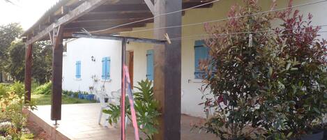 A covered patio area with a wooden pergola, potted plants, and a white building with blue shutters in the background.