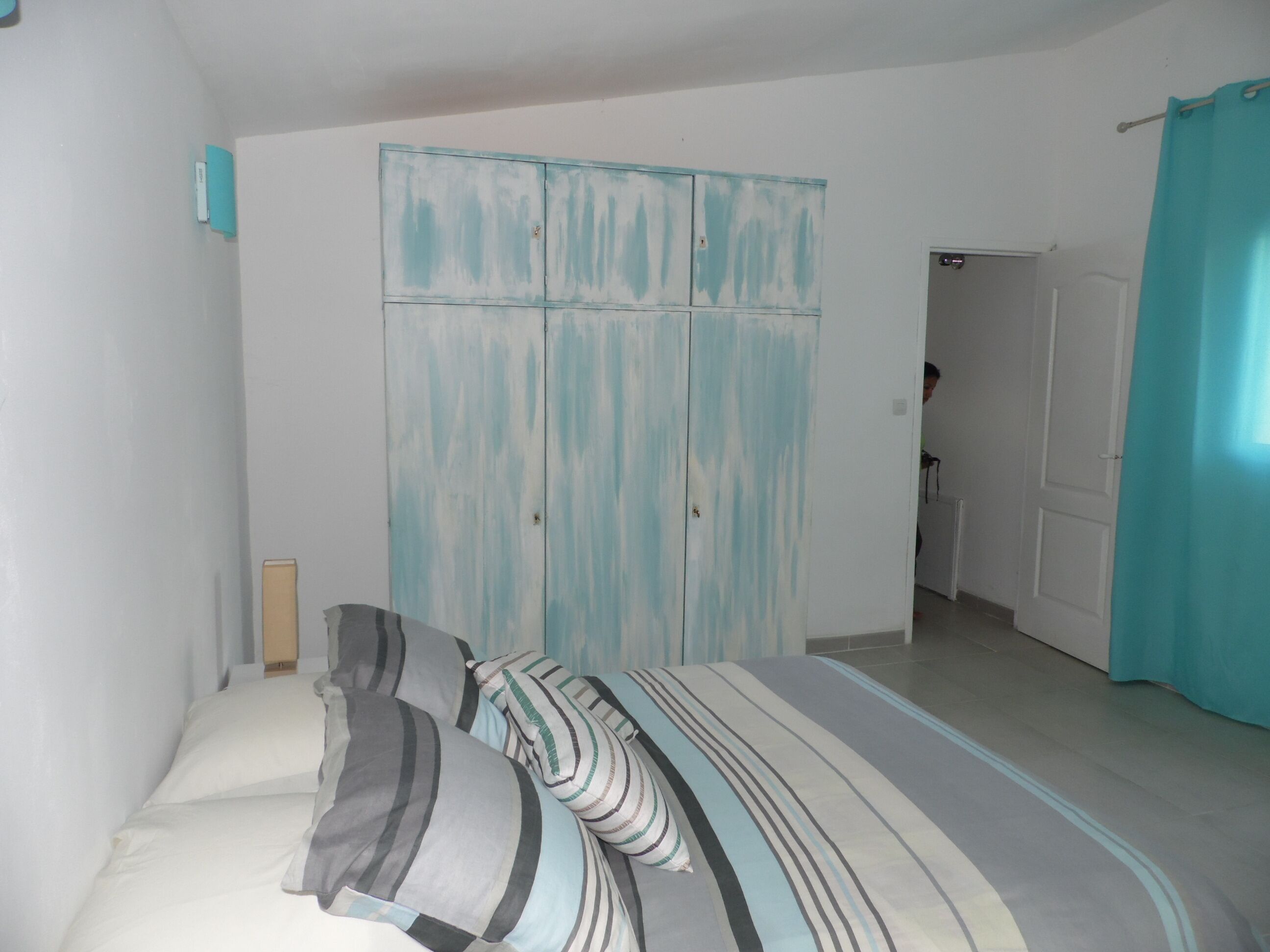 Un dormitori amb un llit, almoines amb ratlles, un armari blau i una porta de bany al fons.