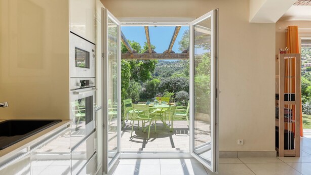 Een moderne keuken met een ingebouwde magnetron en oven, een glazen deur die leidt naar een patio met een tafel en stoelen, en een houten constructie boven de deur.