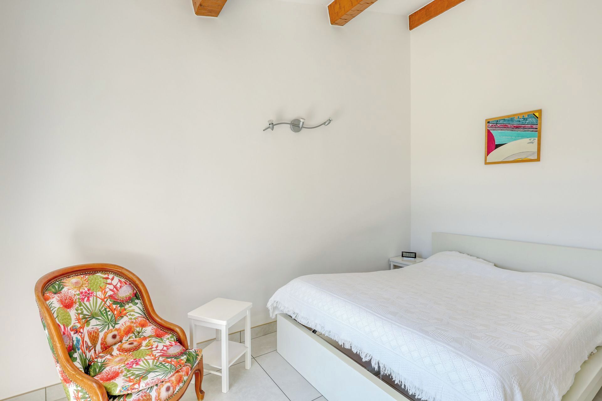 Un dormitorio con una cama, una silla, una mesita pequeña, una lámpara de pared y un cuadro.