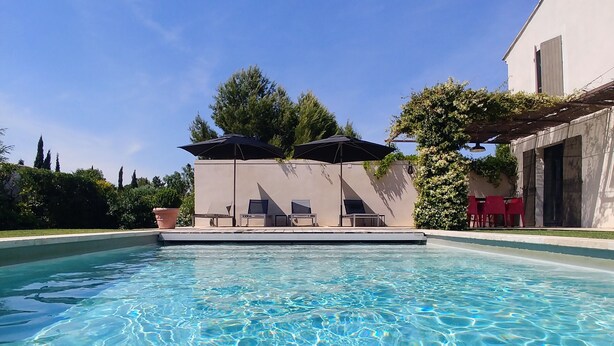 A clear swimming pool with lounge chairs and umbrellas, surrounded by greenery and a building. - Villa, private heated pool, beautiful view of the Alpilles (Eygalières)