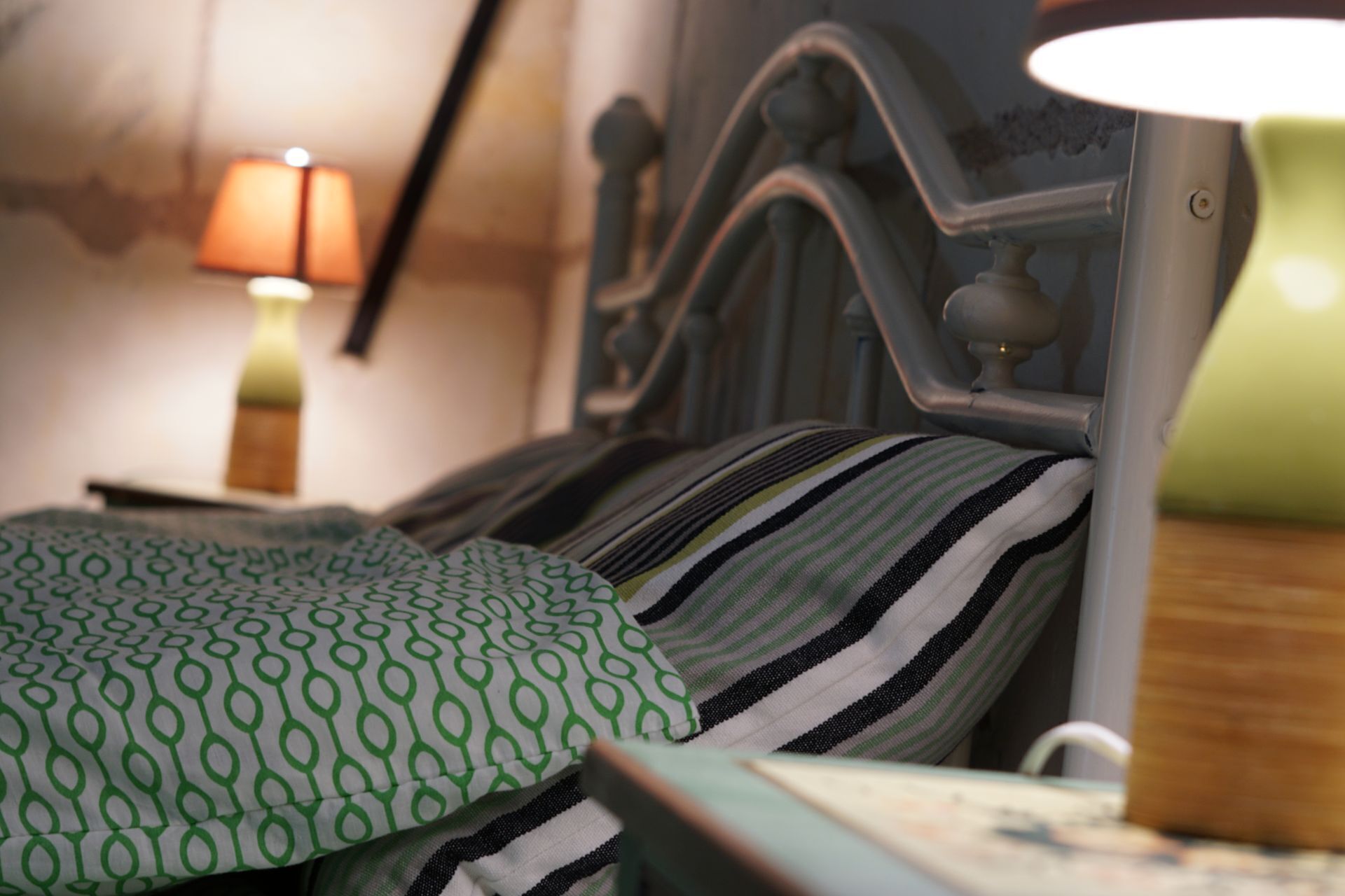 A bed with a striped pillow and a green patterned pillow, a metal headboard, and a bedside lamp.