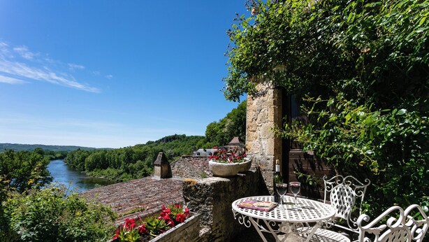 Eine Terrasse mit Tisch und Stühlen, eine Steinmauer und Blick auf Fluss und Wald.