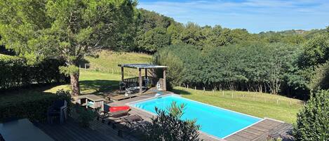 A backyard with a swimming pool, wooden deck, and a covered seating area with chairs and a table.