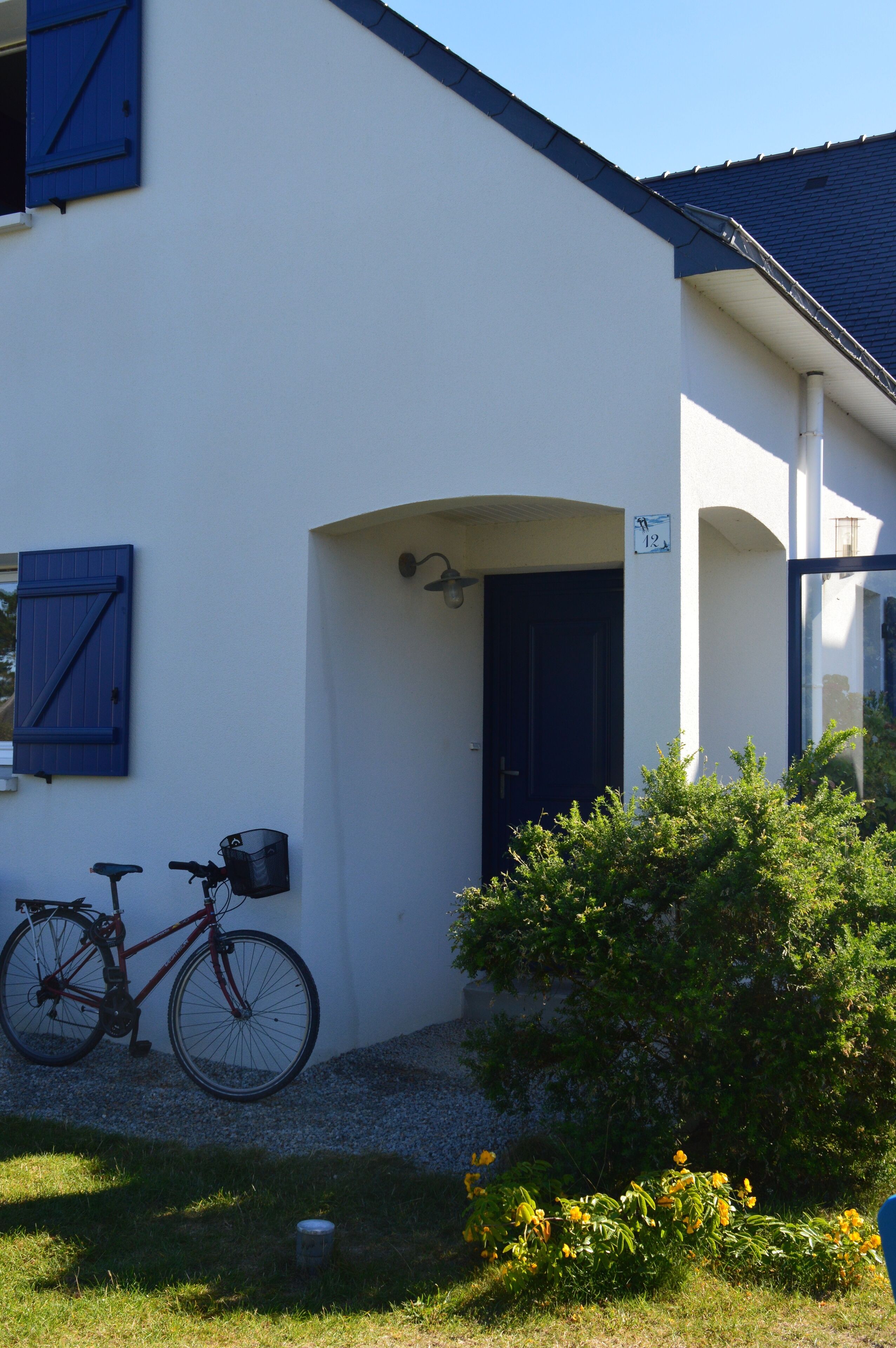 Un bâtiment blanc avec une porte et des volets bleu foncé, un vélo garé devant, et un petit jardin avec des fleurs jaunes.
