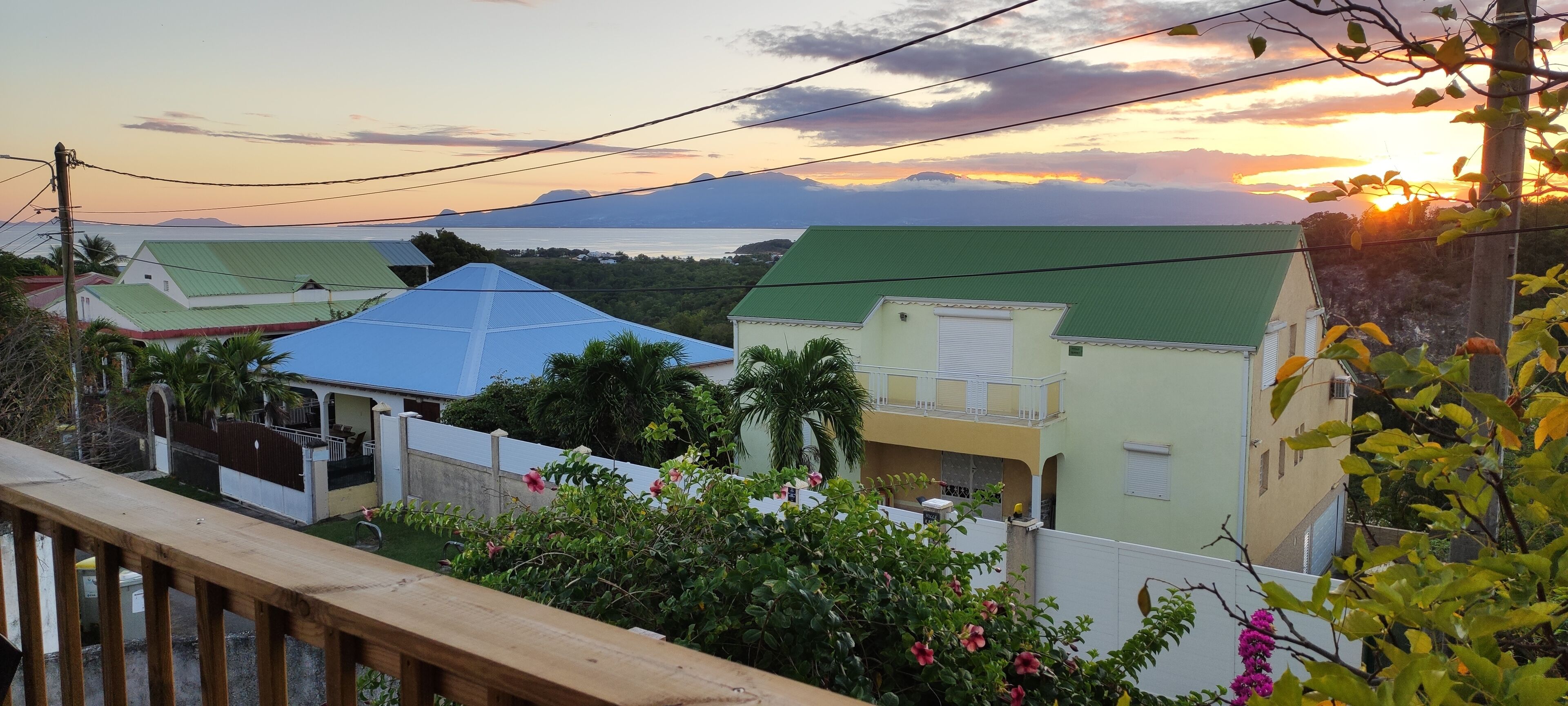 A residential area with houses, a wooden fence, and a view of the sea and mountains during sunset.