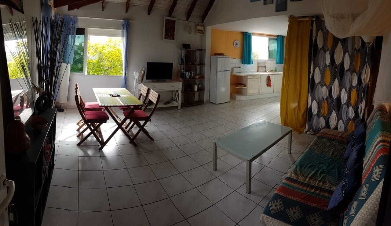 A spacious living area with a dining table and chairs, a sofa, a kitchen area, and a large window with curtains.