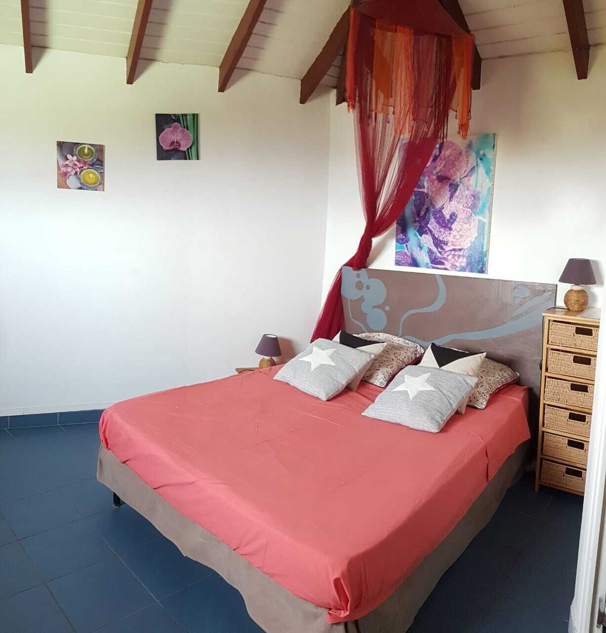 A bedroom with a bed, a nightstand, a lamp, a wicker side table, and a painting on the wall.