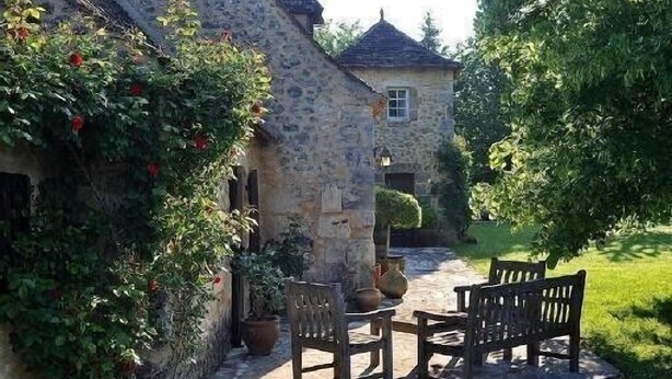 A stone building with a small window and a door, surrounded by greenery and a paved area with wooden furniture. - House 12 Pers. Hamlet Dordogne (Saint-Martin-Des-Combes)