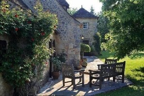 A stone building with a small window and a door, surrounded by greenery and a paved area with wooden furniture. - House 12 Pers. Hamlet Dordogne (Saint-Martin-Des-Combes)