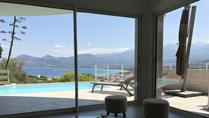 A modern room with a glass table, two ottomans, a green vase, and a view of a pool and mountains. - CALVI, MAGNIFICENT CONTEMPORARY VILLA WITH SEA VIEW AND HEATED POOL  (Calvi)