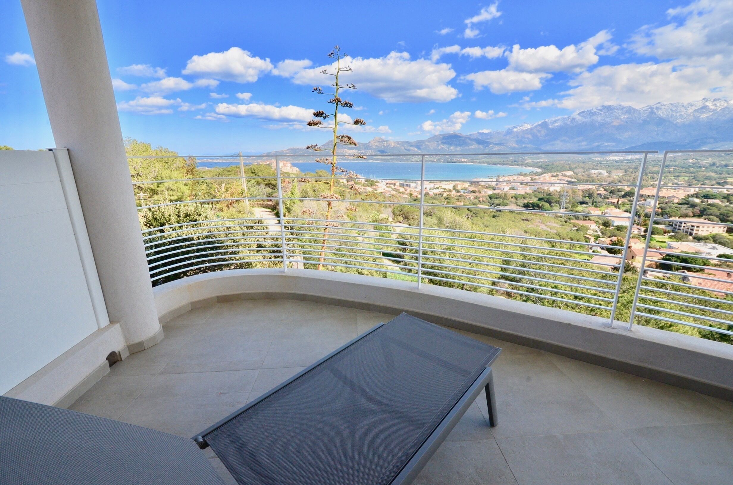 A balcony with a metal railing, a lounge chair, and a view of a mountainous landscape and a body of water.