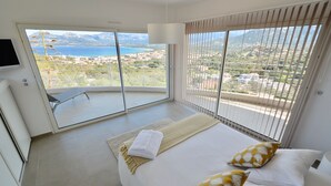 A bedroom with a large bed, two pillows, a bedside table, a TV, and a view of the sea and mountains. - CALVI, MAGNIFICENT CONTEMPORARY VILLA WITH SEA VIEW AND HEATED POOL  (Calvi)