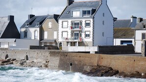 A coastal building with multiple windows and balconies, situated next to a rocky shoreline with waves crashing against a stone wall. - A studio located in a house directly on the beach in a stunning maritime site (Guilvinec)